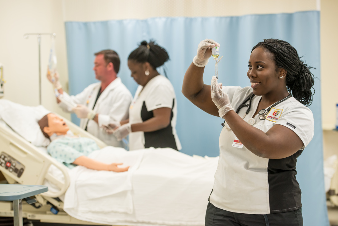 A student practices using a vial as two other students practice bedside nursing skills in an educational lab environment.